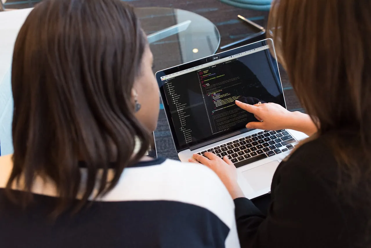 Two women sit at a table, focused on a laptop displaying code for website design and development.  