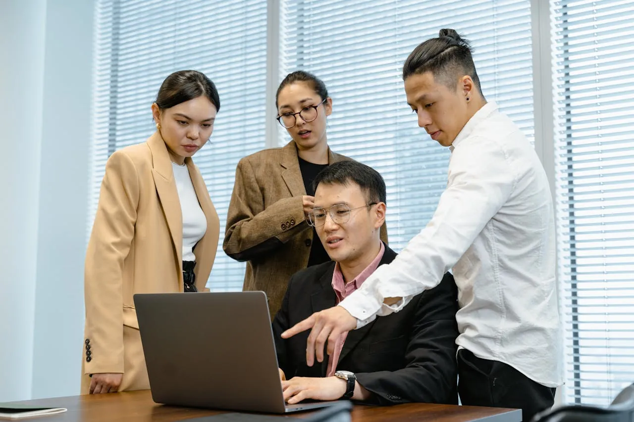 Four digital marketing agency professionals are gathered around a laptop in a bright office. One person is seated, attentively listening, while the others discuss and point at the screen, conveying teamwork and focus. 