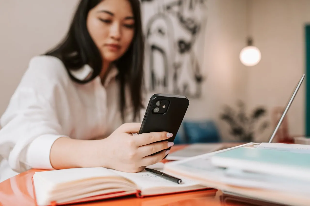A woman in a white blouse, sitting at an orange table, focuses on her smartphone.