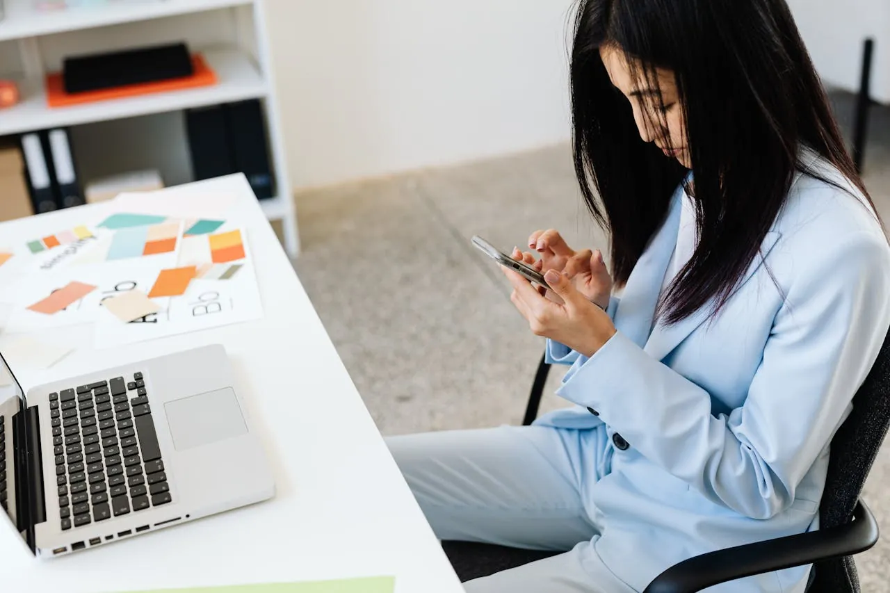 A woman in a light blue suit sits at a white desk, focused on her smartphone. 
