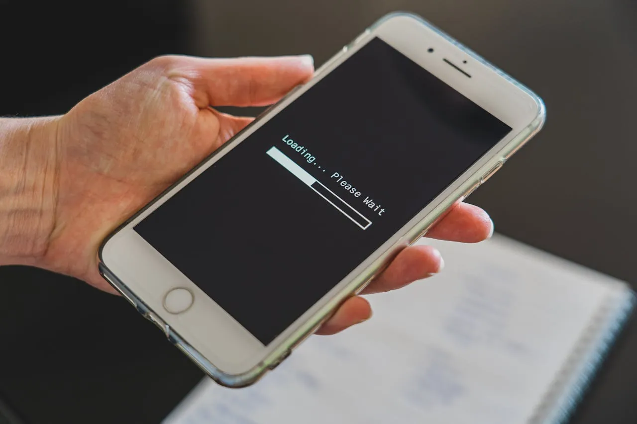 A hand holds a smartphone displaying a "Loading... Please Wait" message with a progress bar, conveying anticipation.