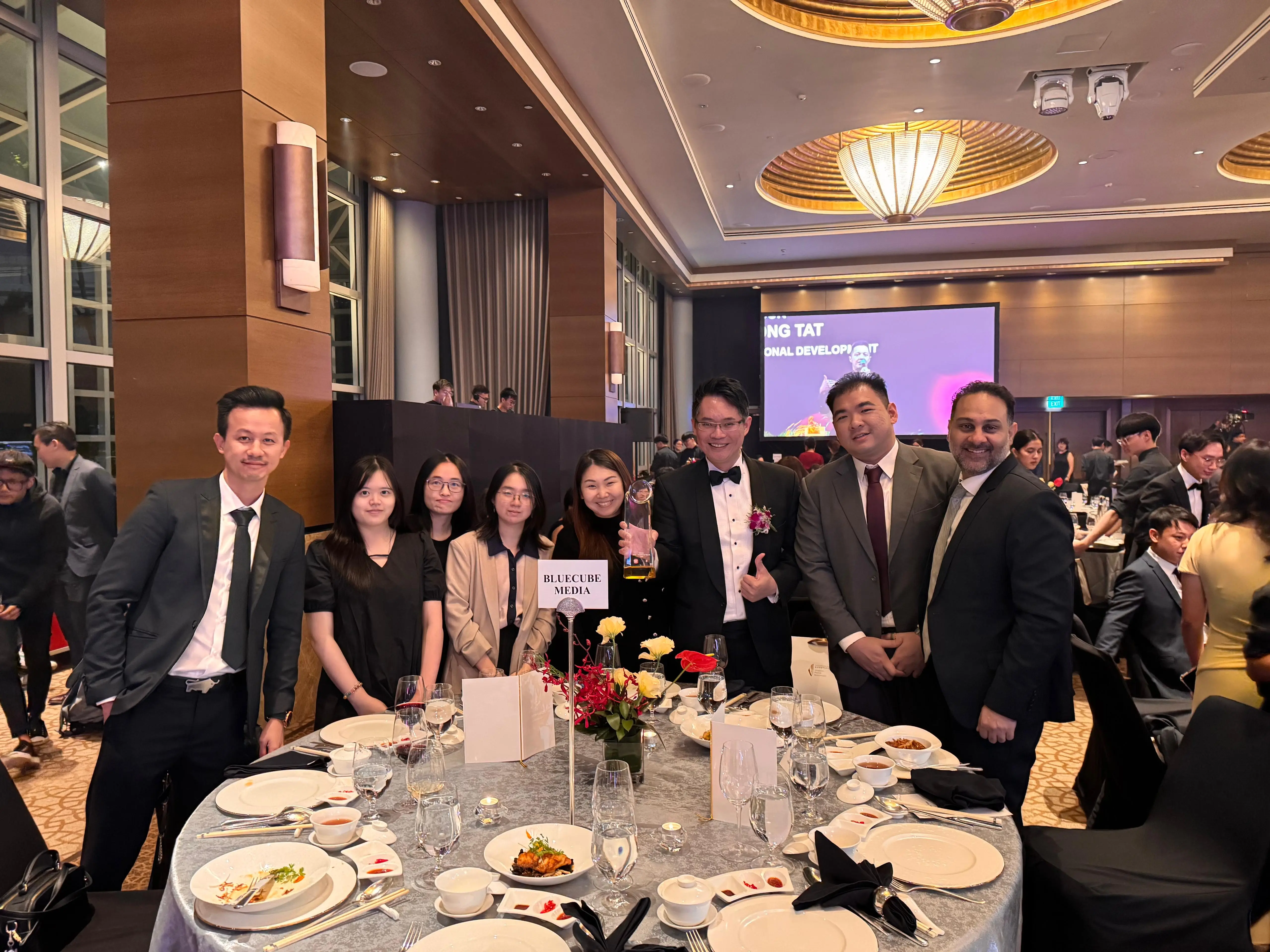 The BlueCube Media team gathered around a gala dinner table, holding the award trophy.