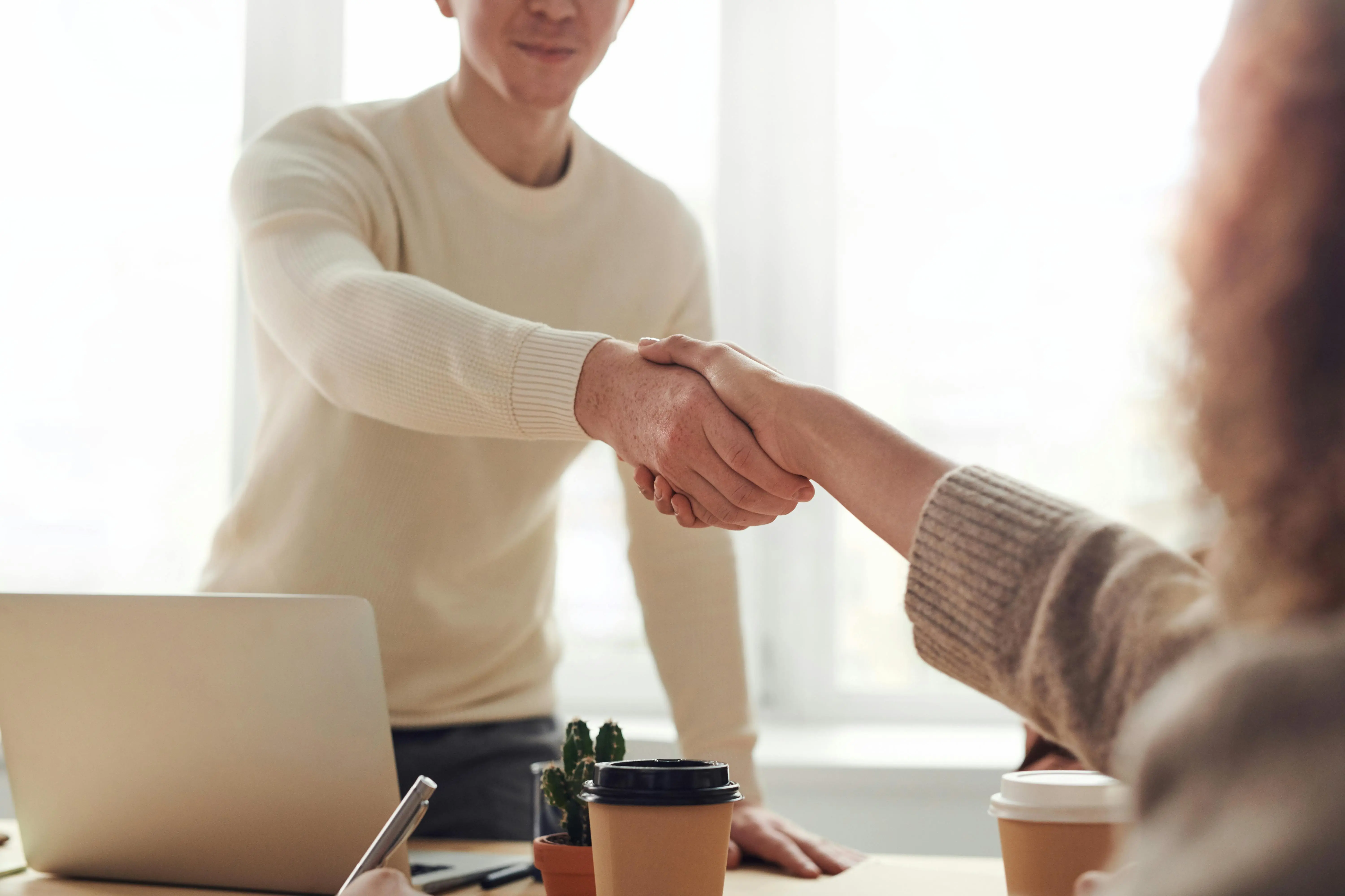 Two people in a cozy meeting setting are shaking hands across a table with laptops and coffee cups, suggesting a successful agreement in a warm atmosphere.