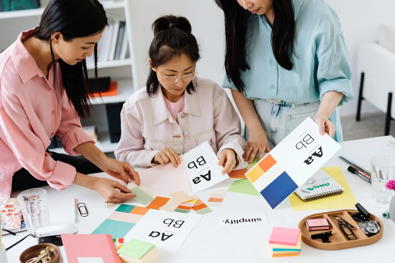 Three people collaborate at a table, examining design samples and color swatches. The atmosphere is focused and creative, suggesting teamwork in a design project. 