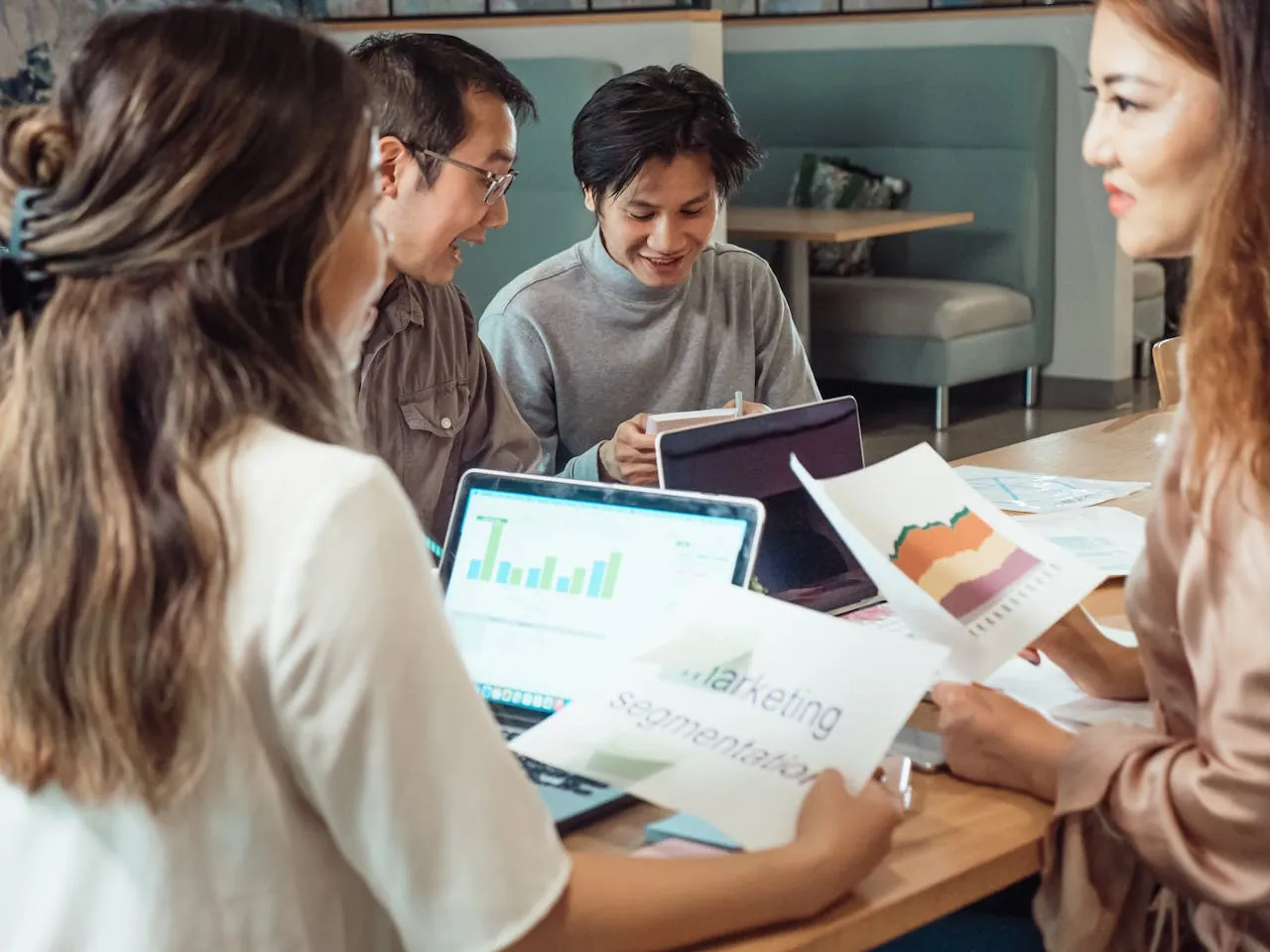 A group of digital marketing agency employees sitting at a table in a modern office. They are discussing marketing data with laptops and graphs. 