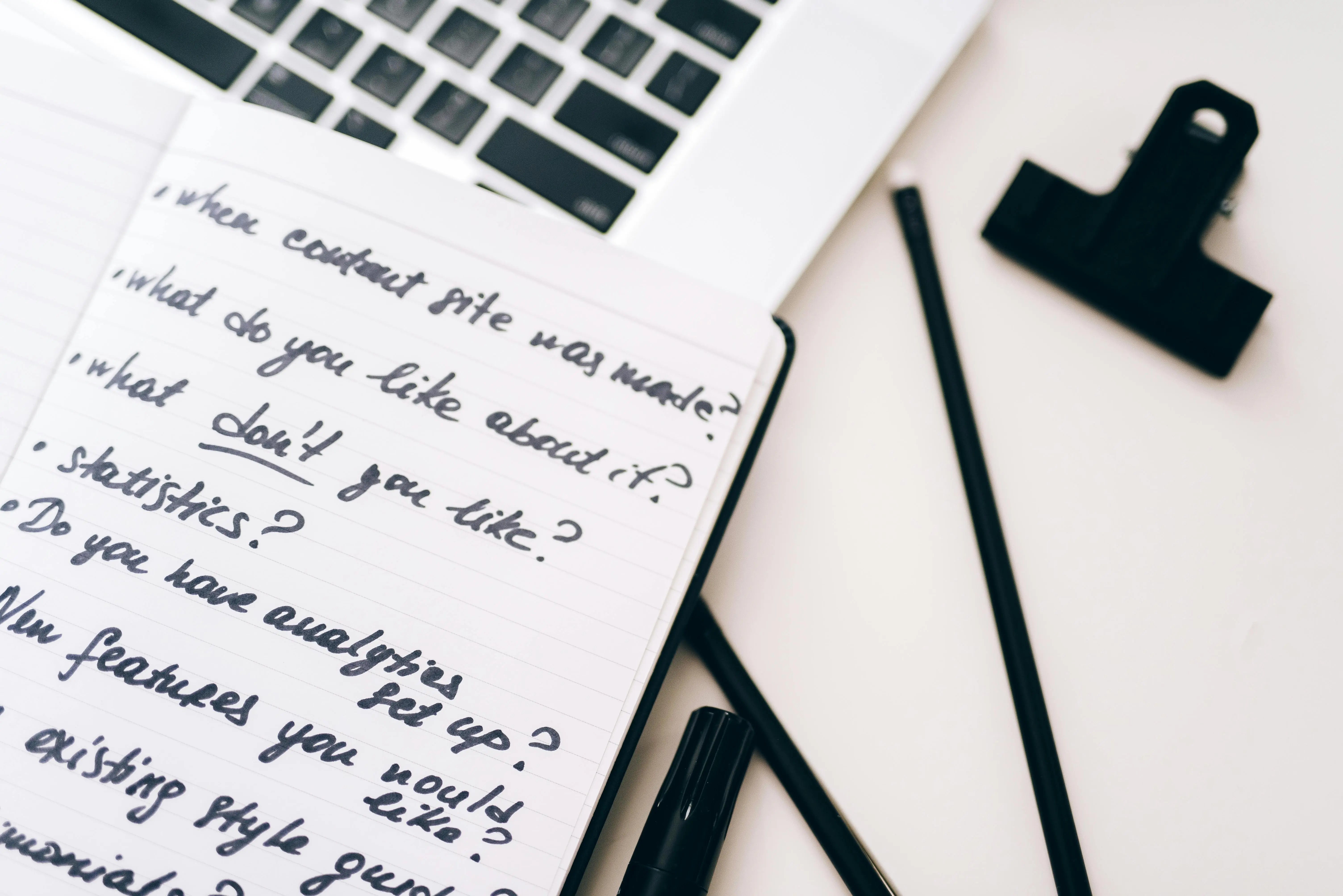 Open notebook with handwritten notes and content strategy questions beside a laptop, black marker, pencil, and a large binder clip on a white desk. 