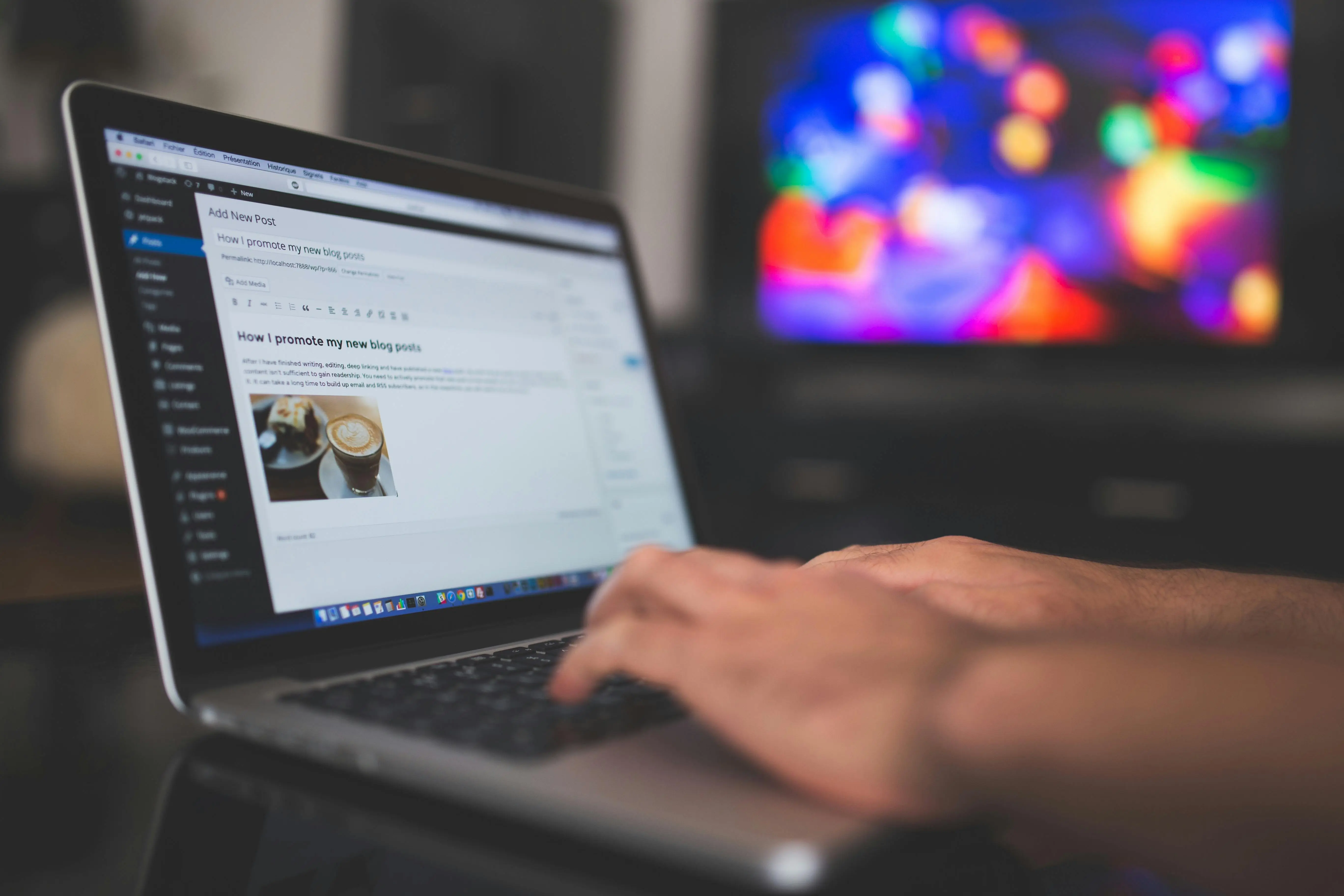 Hands typing on a laptop displaying a blog post editor, with a blurred, colorful TV screen in the background.