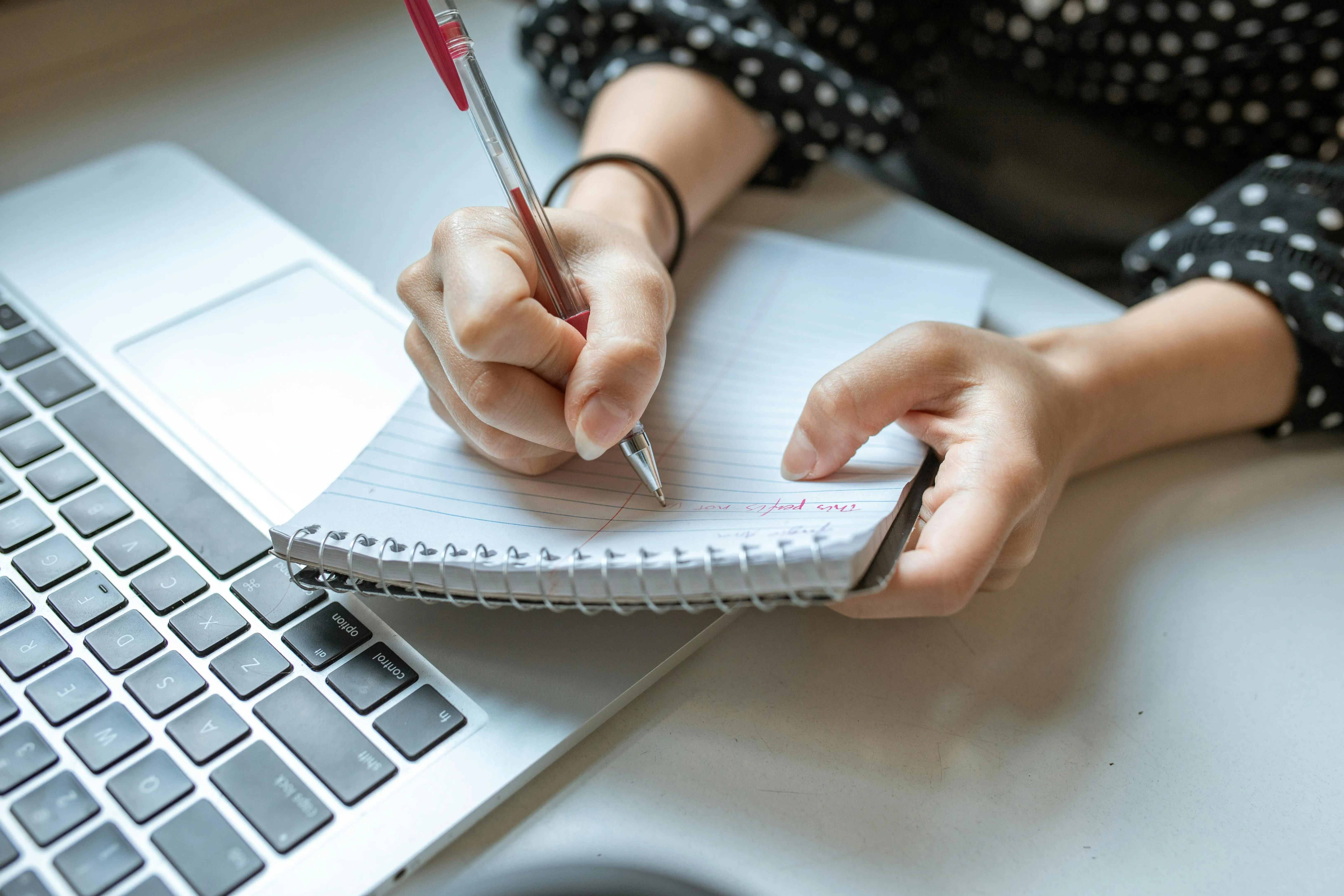 Close-up of a person writing in a spiral notebook with a red pen, next to a laptop. 