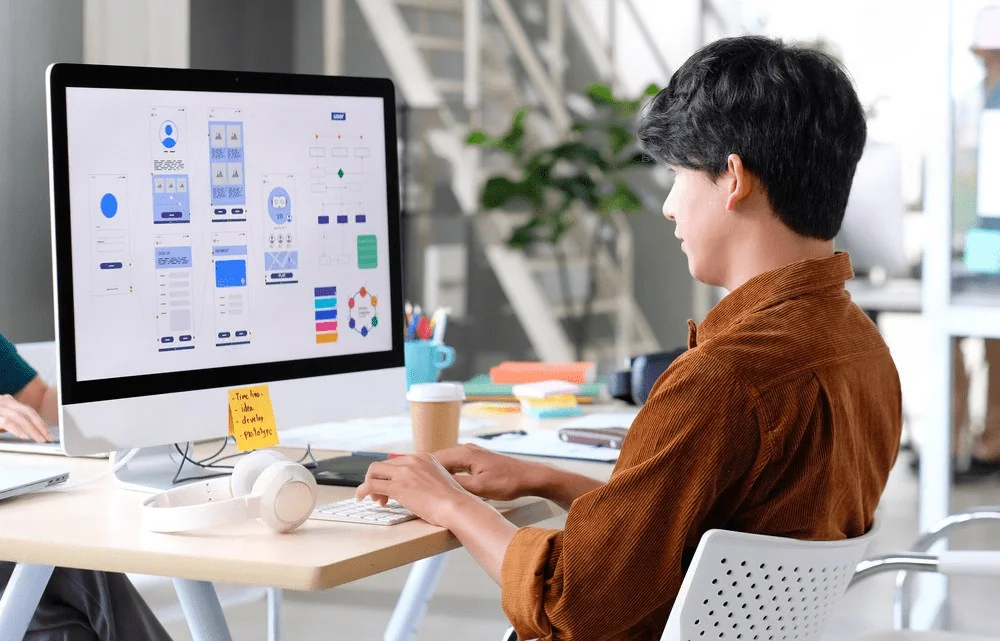 A designer looks at UI/UX layouts of a website on a large screen at his workstation in the office.